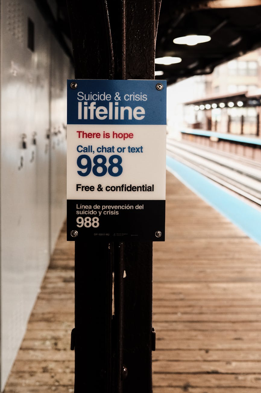 A sign on a train platform reads “Suicide & crisis lifeline” with the number 988 prominently displayed. It encourages people to call, chat, or text for free and confidential support, emphasizing “There is hope.” The platform is empty, suggesting quiet or solitude, but the message is one of connection and help.