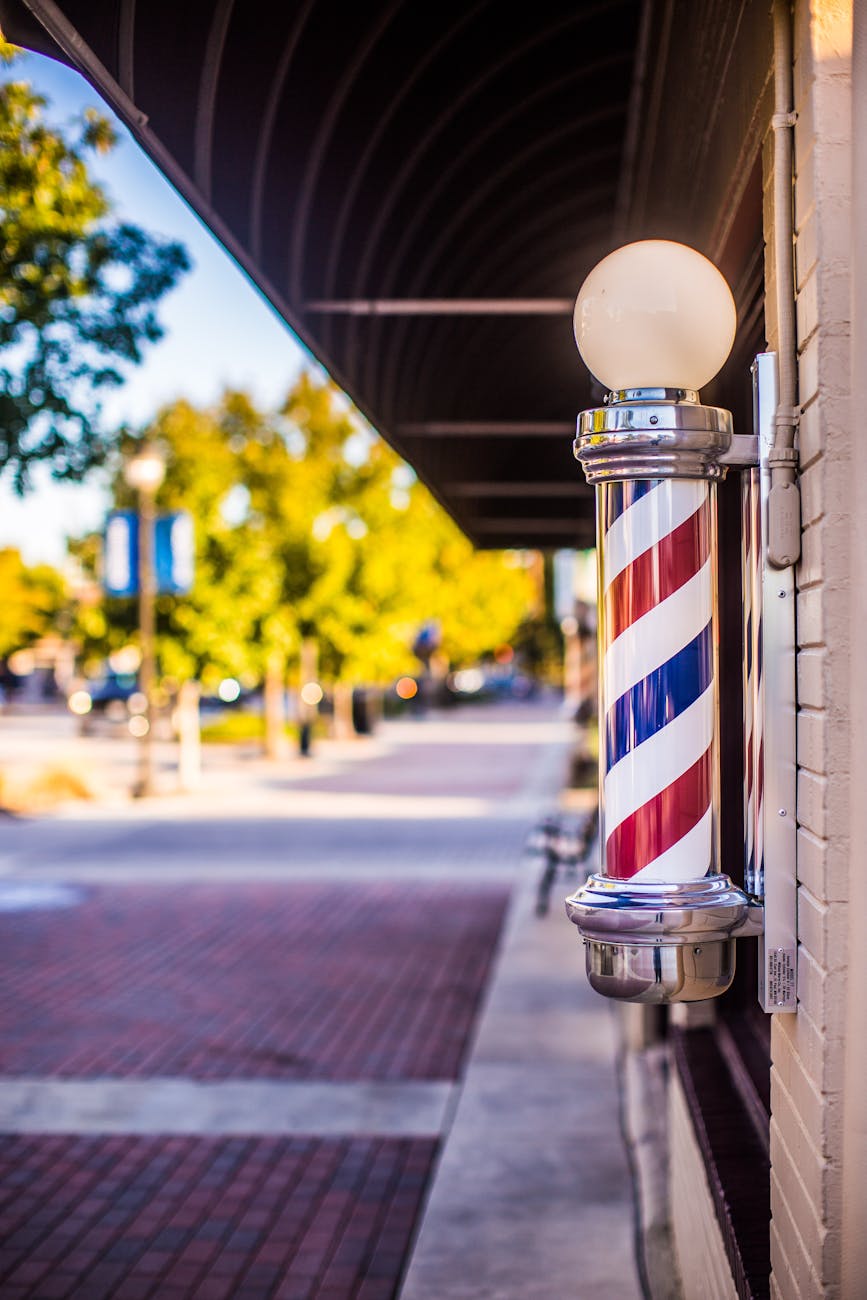 A classic red, white, and blue barber pole mounted on the exterior wall of a barbershop along a quiet sidewalk with trees and storefronts in the background.